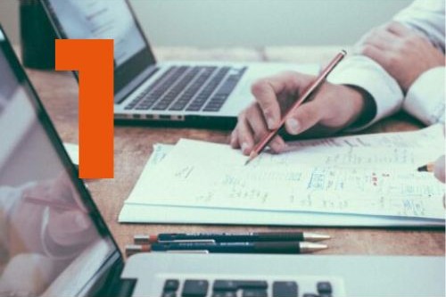 Deux personnes travaillent sur un bureau avec un ordinateur portable ouvert, des documents manuscrits et des crayons, illustrant l’étude des besoins et l’analyse des données.